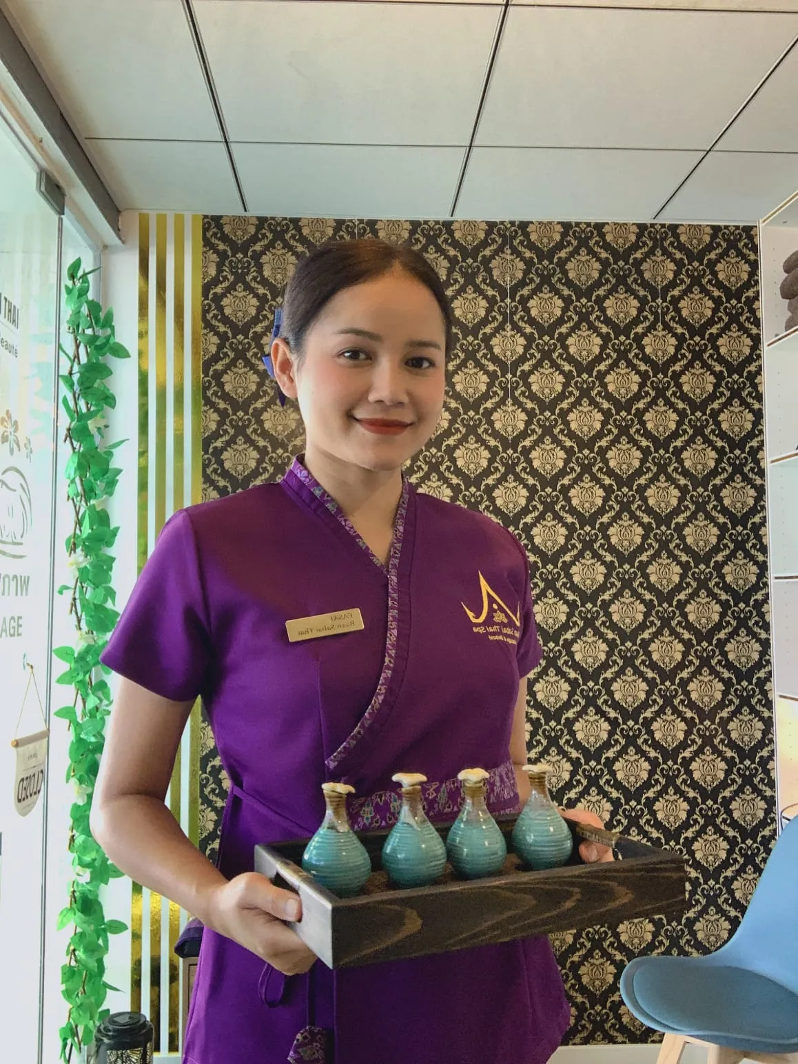 Une praticienne du salon en tenue violette, souriante, un plateau de flacons d'huile entre les mains